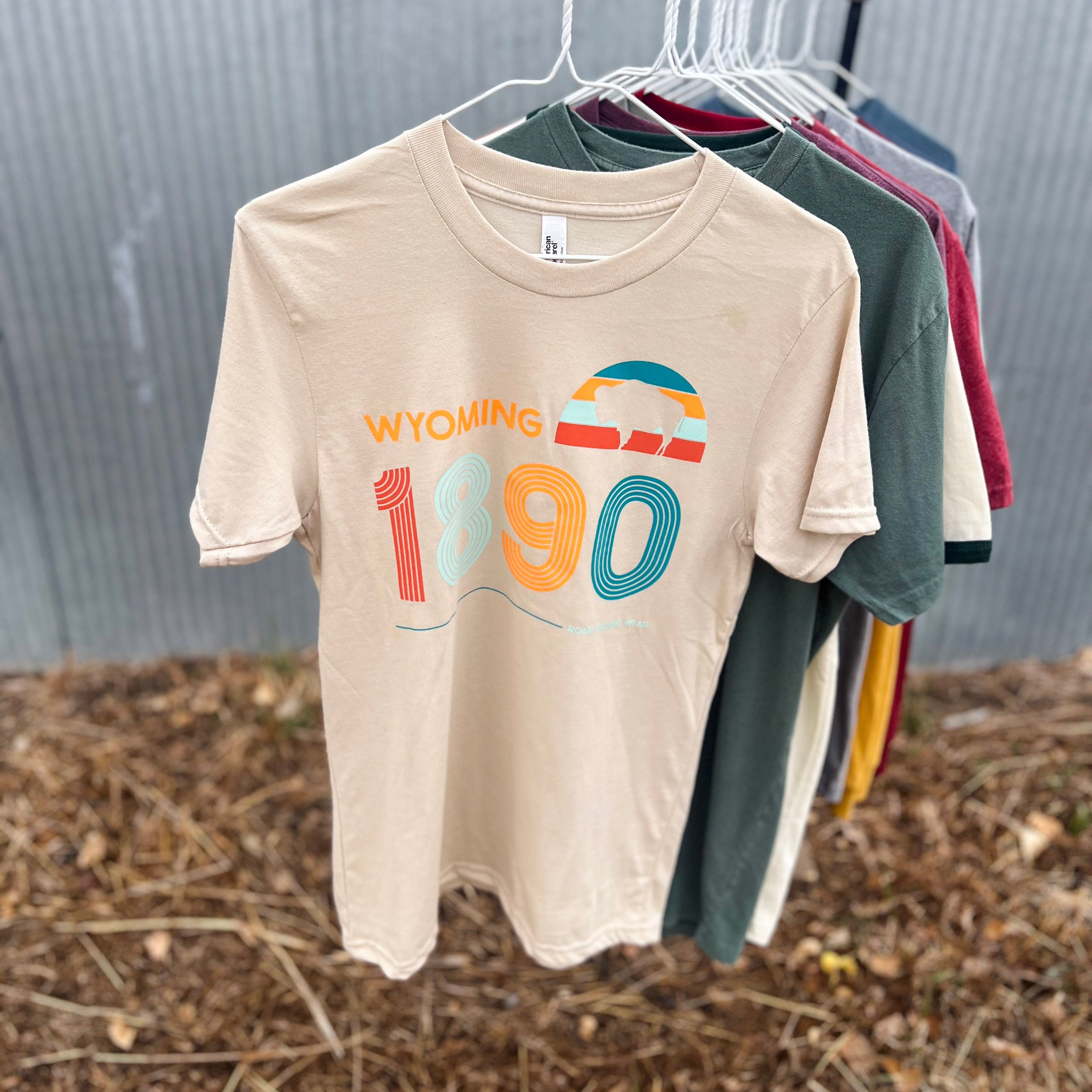 A beige graphic t-shirt hanging on a black clothing rack outdoors, with other shirts in green, red, and blue behind it. The shirt features a retro-style 'WYOMING 1890' graphic in orange and teal.