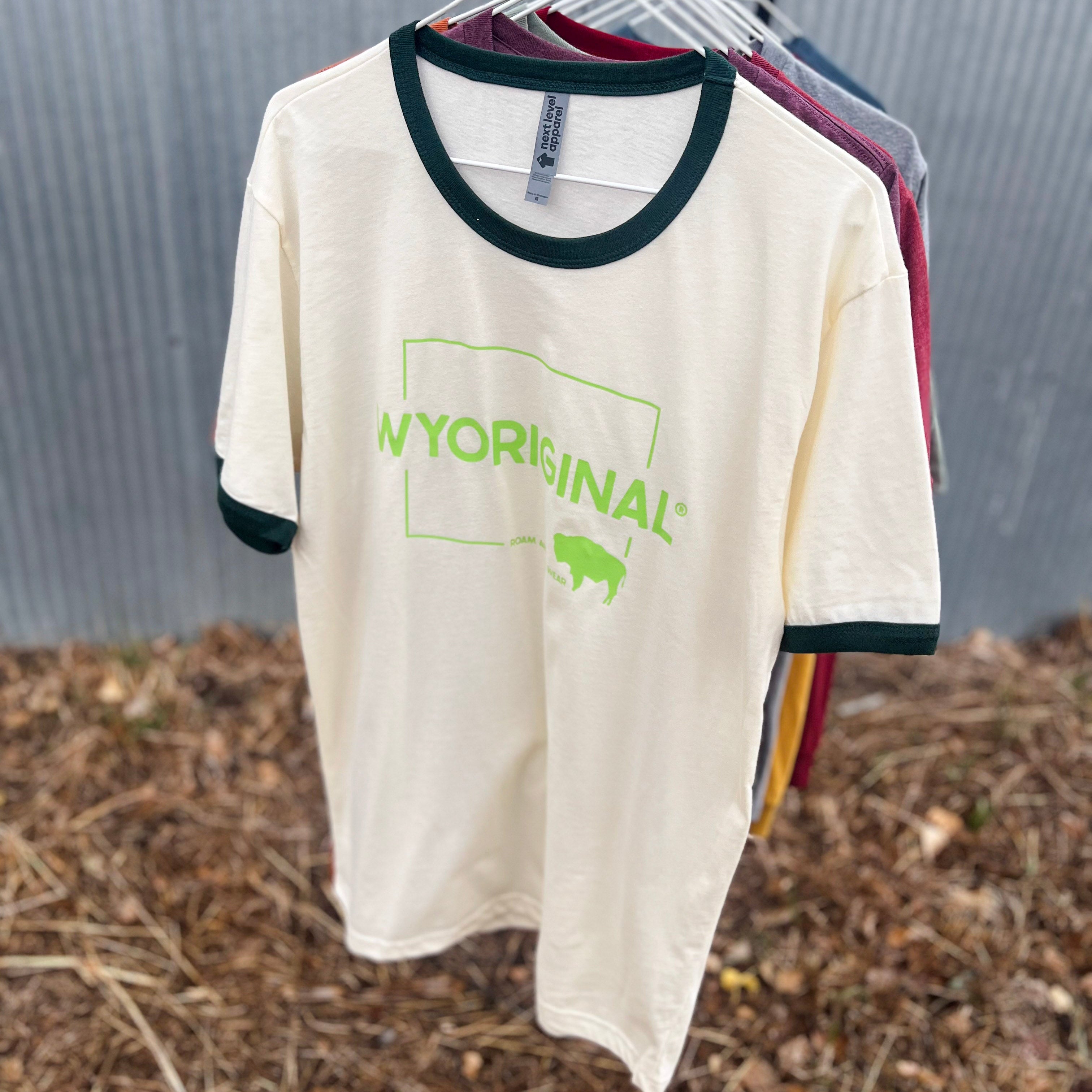 A cream-colored ringer t-shirt with dark green trim and a green 'NYORIGINAL' buffalo graphic, hanging on a black rack outdoors against a corrugated metal wall and wood mulch ground.