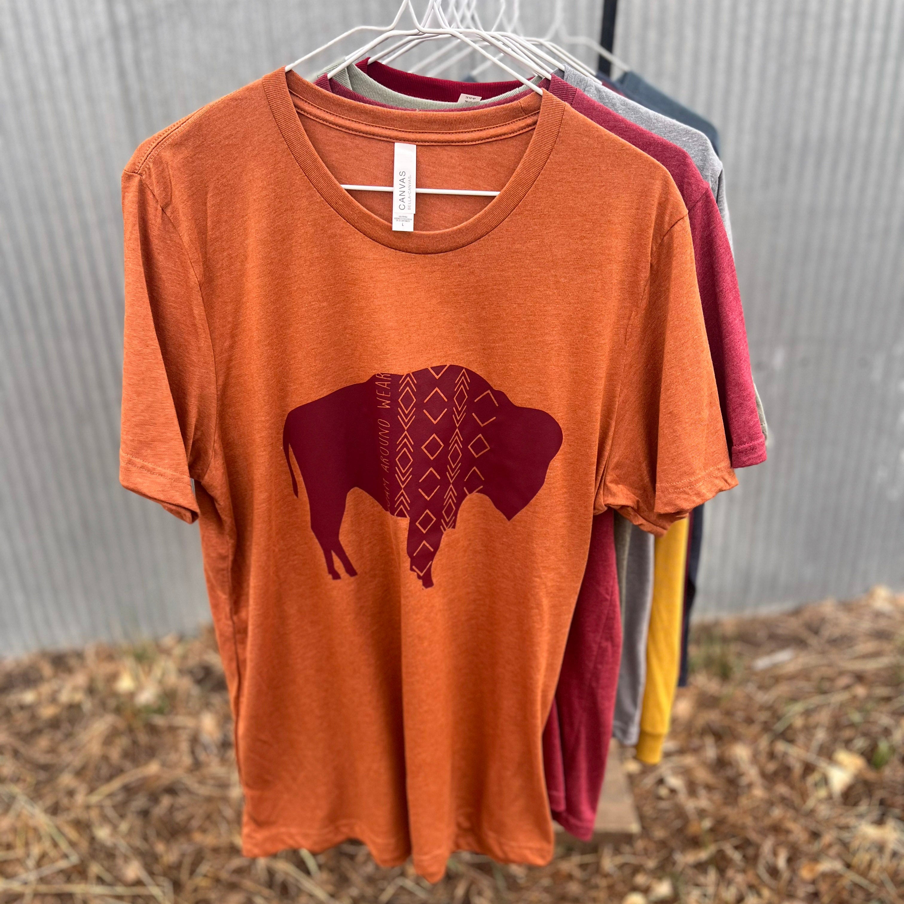 An orange short-sleeve t-shirt hanging on a white hanger, featuring a dark red bison graphic with a geometric diamond pattern on the chest. Other colored t-shirts are visible on hangers behind it against a corrugated metal wall and dry ground background.